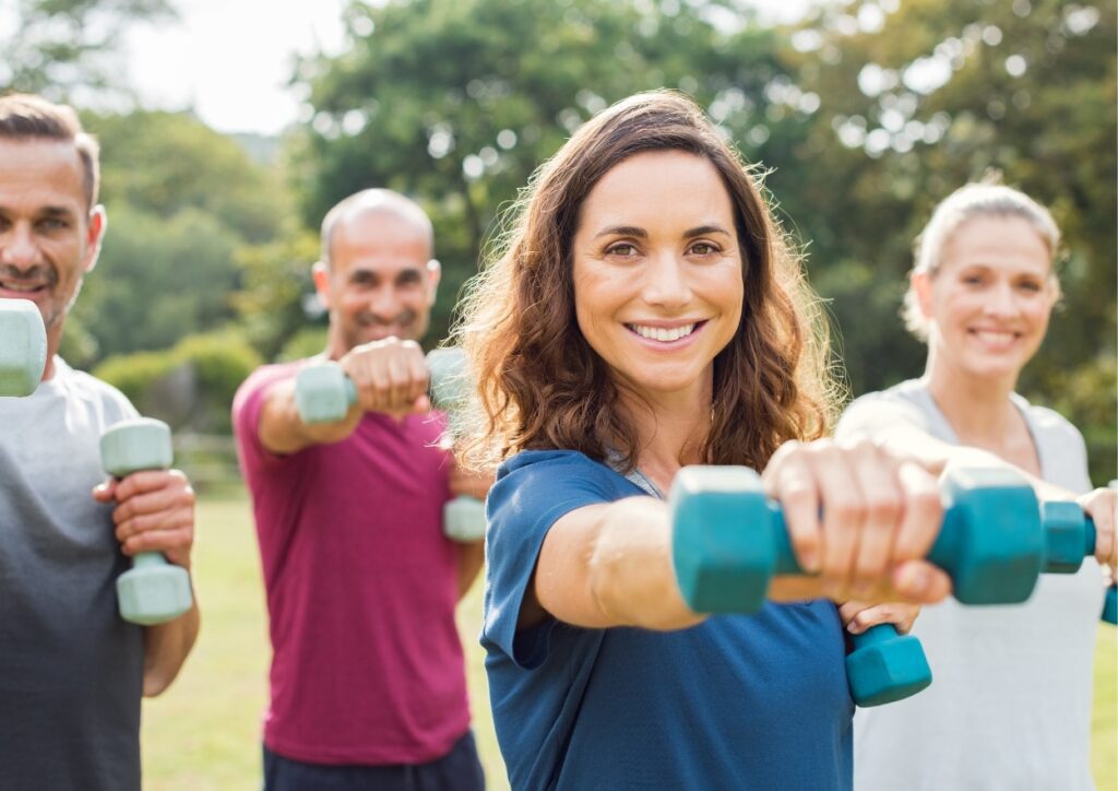 40 plussers trainen buiten in een klein groepje, hebben in 1 hand een dumbell die ze omhoog houden (90 graden)