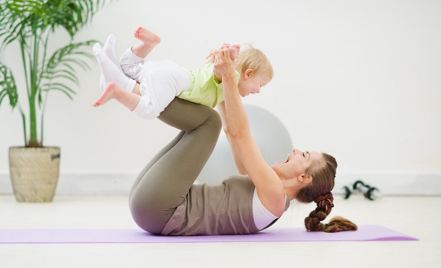 Moeder in sportkleding ligt op een sportmatje in de woonkamer. Moeder tilt haar baby boven zich terwijl zij op haar rug ligt. Moeder en baby lachen