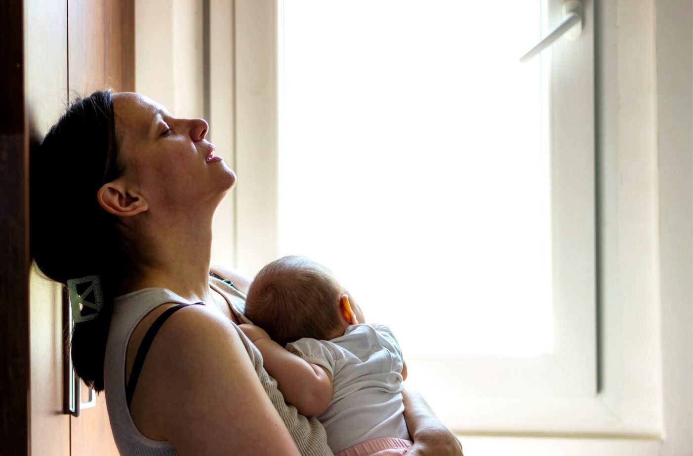 Vermoeide moeder met ogen dicht, leunt achterover tegen een muur, met baby in haar armen