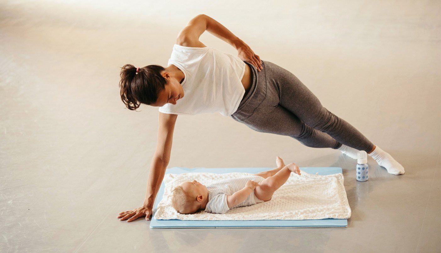 Vrouw in sportkleding traint, terwijl ze lachend kijkt naar haar liggende baby. Vrouw hangt als ware boven de baby, steunend op 1 arm. Andere arm houdt zij in haar zijde.