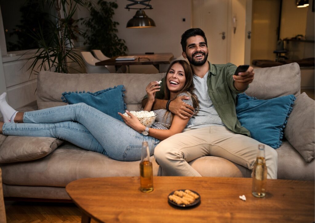 Stel ligt op de bank voor de tv en eten chips. Hebben verder hapjes op tafel staan met ieder een flesje bier.