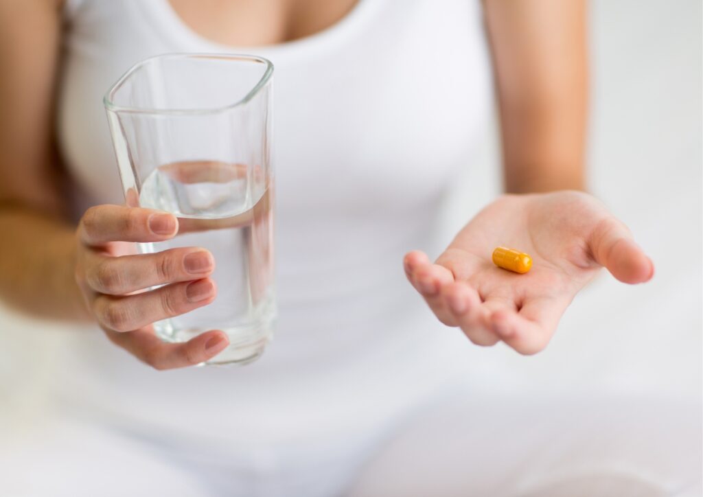 Zittende vrouw in witte kleding houdt een glas water in haar ene hand en een pil in haar andere hand. Hoofd en voeten zijn niet in beeld.
