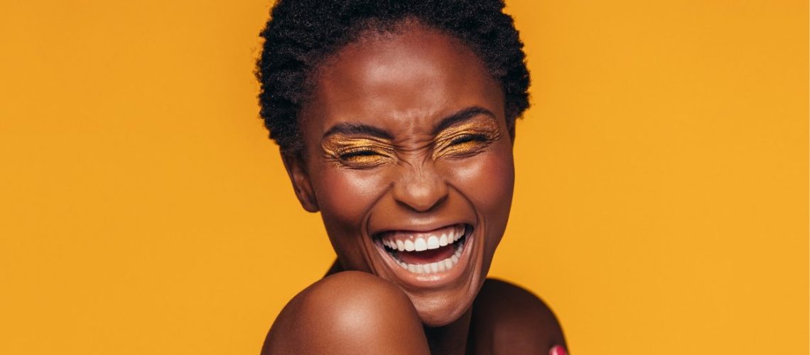 Vrolijke vrouw lacht met haar ogen dicht. Heeft een donkere huid en felgele oogschaduw. Staat voor een fel oranje, gele achtergrond.
