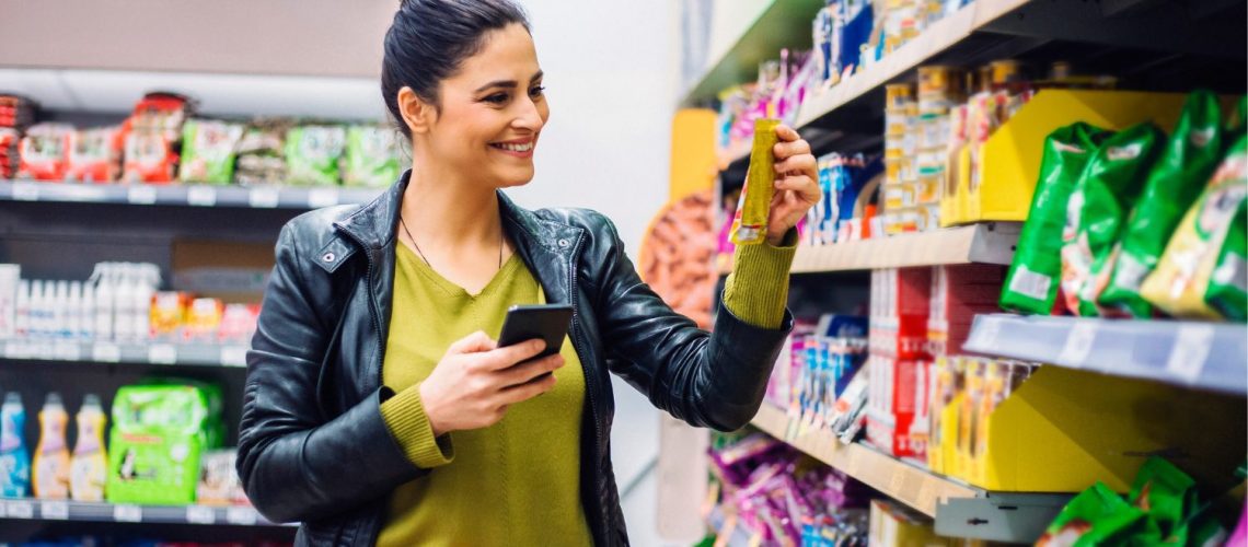 Vrouw met boodschappenwagen doet boodschappen in de supermarkt en bekijkt lachend een product die zij vasthoudt in haar hand.