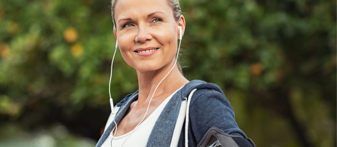 50+ vrouw kijkt lachend, net langs de camera. Draagt sportkleding en heeft airpods in. Telefoon zit in een hoesje aan haar arm. Houdt haar handen in de zij.