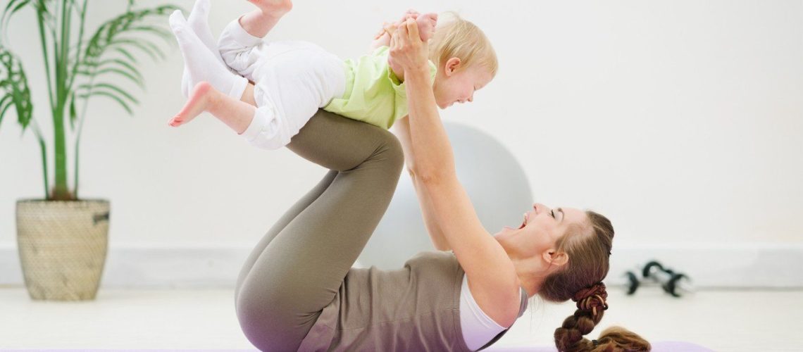 Moeder in sportkleding ligt op een sportmatje in de woonkamer. Moeder tilt haar baby boven zich terwijl zij op haar rug ligt. Moeder en baby lachen