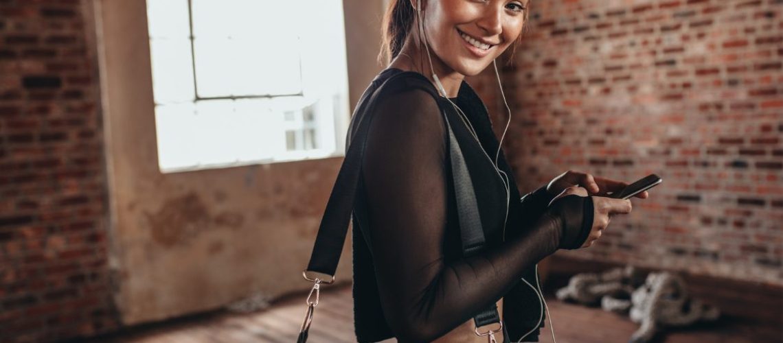 Vrouw in sportkleding met sporttas om schouder, lacht in camera. Ze heeft haar telefoon vast en heeft oordopjes met muziek in haar oren
