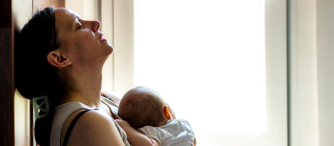 Vermoeide moeder met ogen dicht, leunt achterover tegen een muur, met baby in haar armen